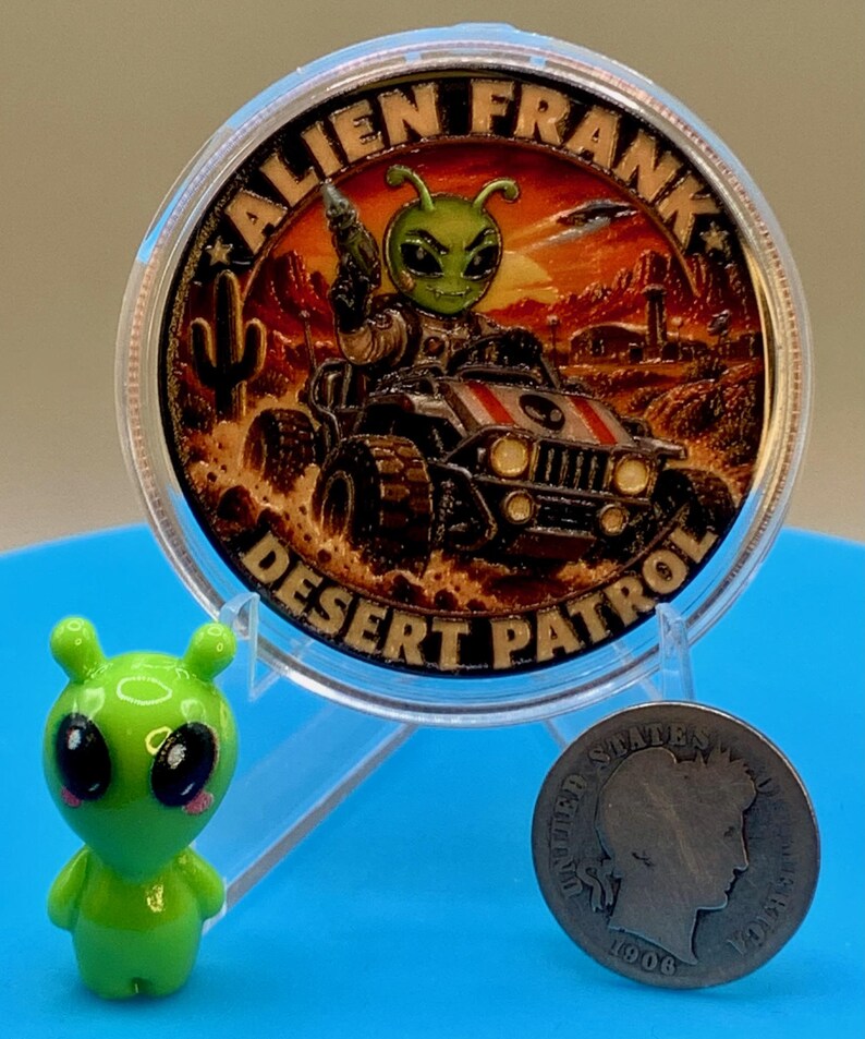 Alien Frank Desert Patrol 1 oz copper round in a clear capsule on a blue stand, shown next to a small green alien figurine and an older silver-colored coin to give a sense of scale.