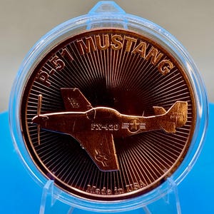 Close-up front view of a 1 oz .999 fine copper round with a raised P-51 Mustang WWII fighter plane design, engraved "FX-420," and "Made in USA," standing in a display holder on a blue background.