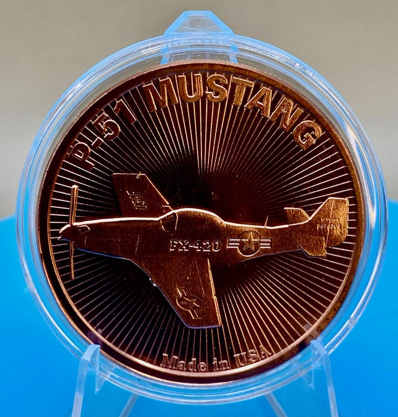 Close-up front view of a 1 oz .999 fine copper round with a raised P-51 Mustang WWII fighter plane design, engraved "FX-420," and "Made in USA," standing in a display holder on a blue background.
