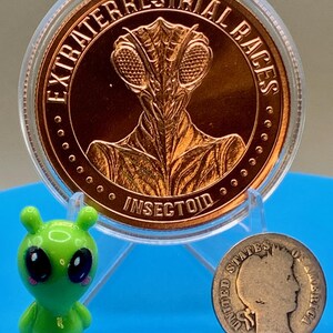 Obverse of the Intaglio Mint Insectoid 1 oz copper round showing a mantis-style alien head and the legend EXTRATERRESTRIAL RACES, shown beside a small green alien figurine and a vintage silver coin for size comparison.