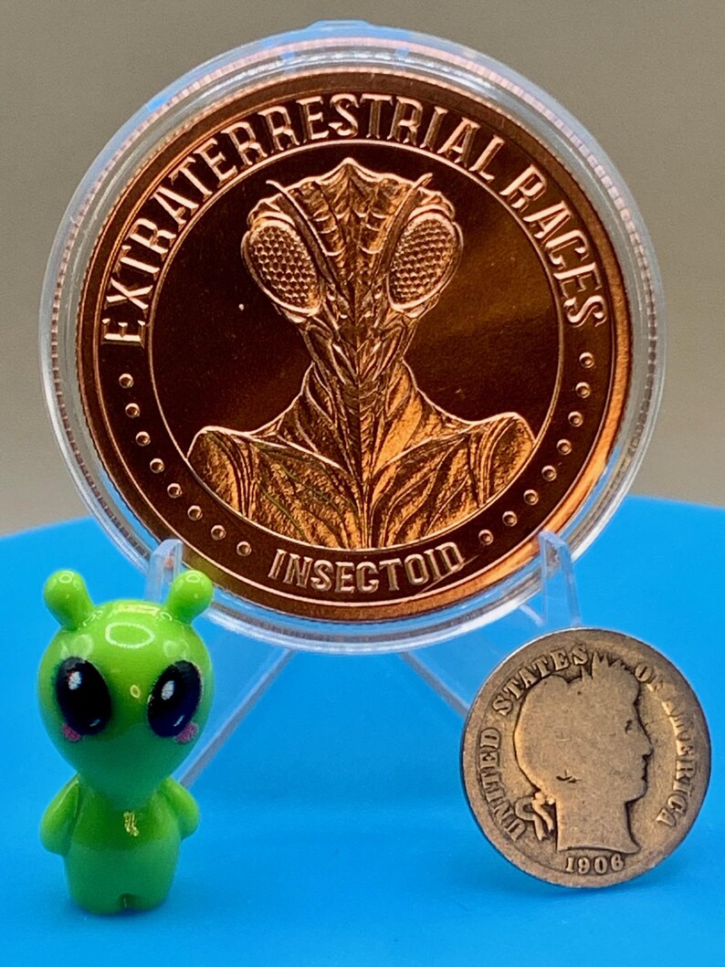 Obverse of the Intaglio Mint Insectoid 1 oz copper round showing a mantis-style alien head and the legend EXTRATERRESTRIAL RACES, shown beside a small green alien figurine and a vintage silver coin for size comparison.