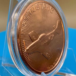 Tilted front view of a 1 oz .999 fine copper round in a display capsule, showing the P-51 Mustang design and round edge at an angle, on a blue background.