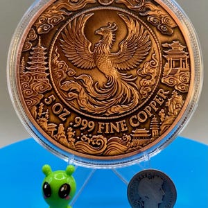Copper Phoenix round in its capsule propped on a stand, shown next to a small green alien figurine and a dime for size comparison, Area51 Precious Metals themed display.