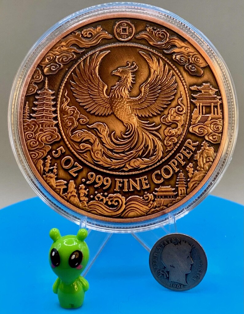 Copper Phoenix round in its capsule propped on a stand, shown next to a small green alien figurine and a dime for size comparison, Area51 Precious Metals themed display.