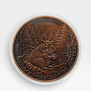 May include: A round copper coin featuring a raccoon sitting in a forest setting. The text reads LIMITED MINTAGE 5000, FIVE AVD P OUNCES, and .999 FINE COPPER. The coin is encased in a clear protective circular capsule against a plain white background.