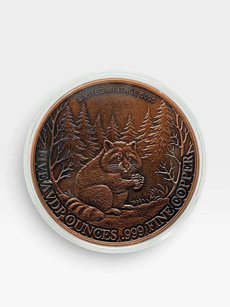 May include: A round copper coin featuring a raccoon sitting in a forest setting. The text reads LIMITED MINTAGE 5000, FIVE AVD P OUNCES, and .999 FINE COPPER. The coin is encased in a clear protective circular capsule against a plain white background.