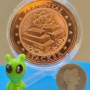 "Elemental Stacker" reverse of the Night Arrival 1 oz copper round in its capsule next to a small green alien figurine and a silver coin for size comparison