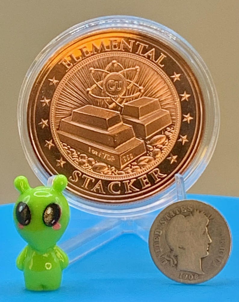"Elemental Stacker" reverse of the Night Arrival 1 oz copper round in its capsule next to a small green alien figurine and a silver coin for size comparison