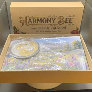Open Harmony Bee copper round packaging box showing the round nested in its capsule on top of a printed certificate insert, with the box lid visible in the background.
