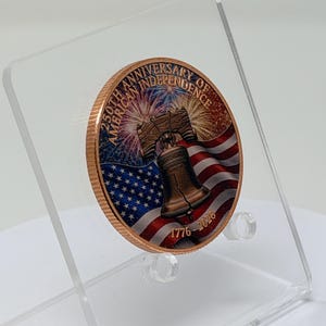 Three-quarter angle view of the colorized Liberty Bell 1 oz copper round on a clear acrylic stand, showing the American flag and fireworks design turned to the side
