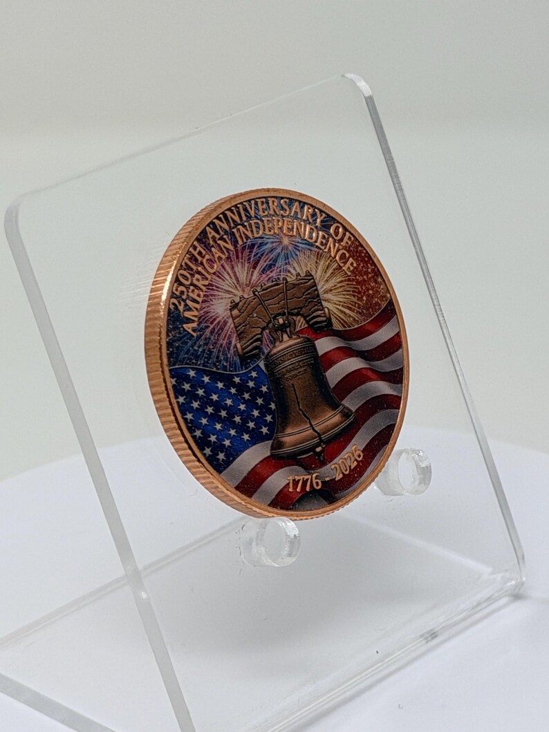 Three-quarter angle view of the colorized Liberty Bell 1 oz copper round on a clear acrylic stand, showing the American flag and fireworks design turned to the side