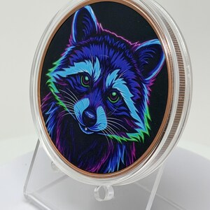 May include: A circular collectible coin encased in a clear protective plastic capsule, featuring a vibrant, neon-colored illustration of a raccoon against a black background, with shades of electric blue, purple, and green highlighting its fur.