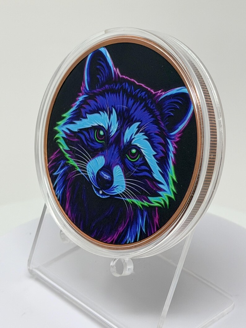 May include: A circular collectible coin encased in a clear protective plastic capsule, featuring a vibrant, neon-colored illustration of a raccoon against a black background, with shades of electric blue, purple, and green highlighting its fur.