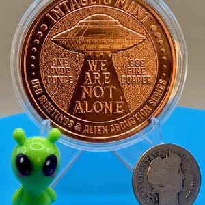 Reverse of the Intaglio Mint UFO Sightings 1 oz copper round in its capsule, shown beside a small green alien figurine and a vintage silver coin to give a sense of the round's size.