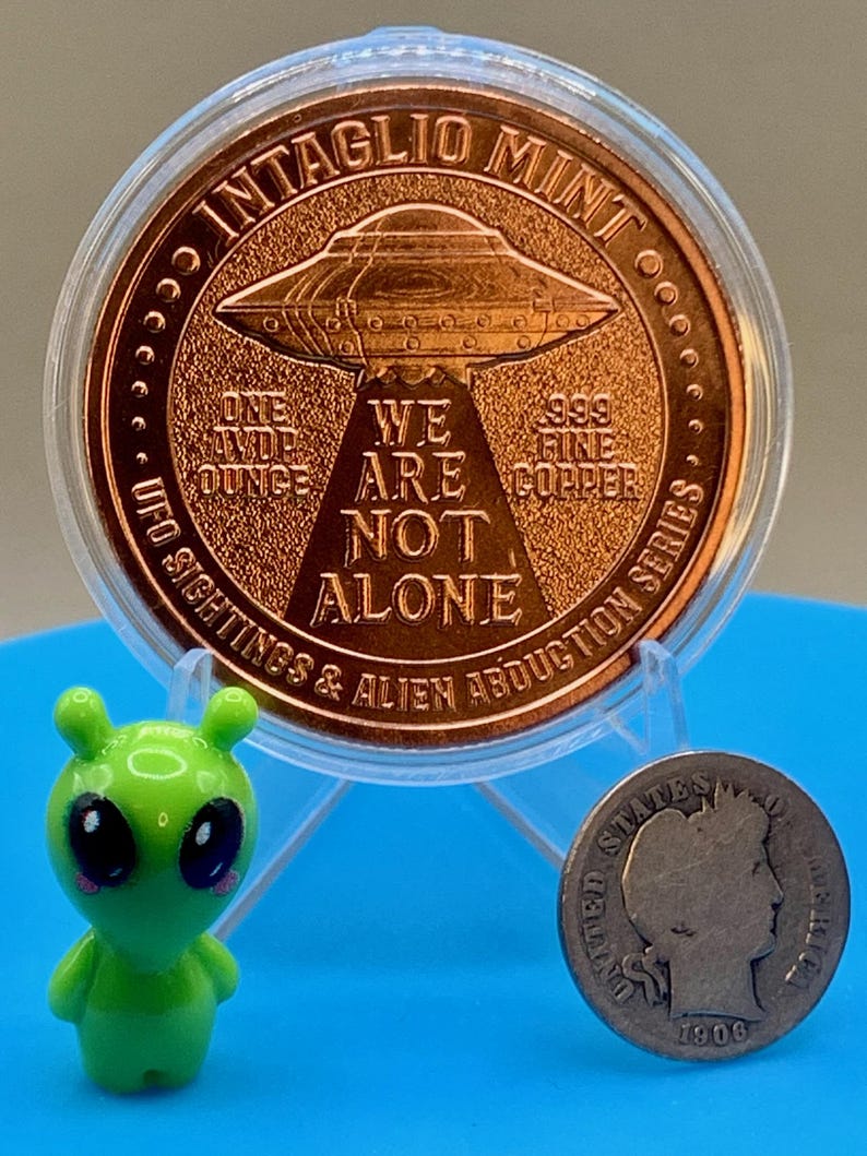 Reverse of the Intaglio Mint UFO Sightings 1 oz copper round in its capsule, shown beside a small green alien figurine and a vintage silver coin to give a sense of the round's size.
