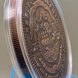 Copper-toned legal tender coin encased in a clear plastic protective capsule, featuring the Republic of Liberia seal, a ship, "The Love of Liberty Brought Us Here," "2026," and "5 AVDP OZ One Dollar" with a reeded edge and metallic finish