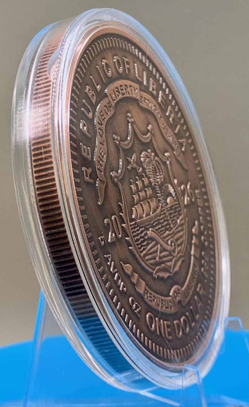 Copper-toned legal tender coin encased in a clear plastic protective capsule, featuring the Republic of Liberia seal, a ship, "The Love of Liberty Brought Us Here," "2026," and "5 AVDP OZ One Dollar" with a reeded edge and metallic finish