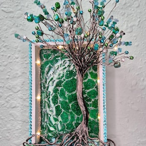 May include: Handmade wire tree sculpture with a green mosaic background. The tree features a twisted wire trunk and branches adorned with green, blue, and clear beads. The frame is white with a blue beaded border and small lights.