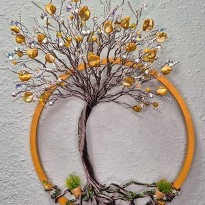 Puede incluir: Un adorno de pared que presenta una escultura de árbol de alambre dentro de un aro de madera circular. El árbol tiene ramas de alambre trenzado marrón oscuro adornadas con cuentas de color ámbar, cristales transparentes y detalles plateados. Dos pequeñas macetas de terracota con follaje verde se sitúan en la base de las raíces. El texto "Handmade With Love by Lisa O'Callaghan" es visible en la esquina superior derecha.