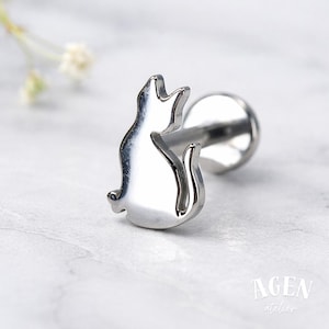 May include: A silver-colored labret stud featuring a stylized cat silhouette. The cat is sitting with its tail curled, and the stud has a flat back for comfortable wear. The jewelry is designed for body piercings.