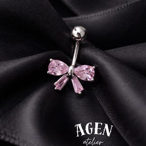 May include: A silver belly button ring with a bow design. The bow features pink teardrop and rectangular-cut gemstones. The jewelry is set against a black satin background. The text "AGEN atelier" is visible.