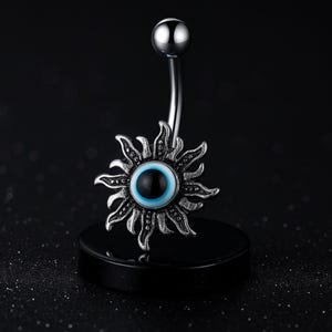 Könnte beinhalten: Ein silberner Bauchnabelpiercing mit einem Sonnenmuster und einem zentralen bösen Blick. Das Auge ist blau und schwarz. Der Ring wird auf einem schwarzen Ständer vor dunklem Hintergrund präsentiert. Der Text "@Agen Atelier" ist sichtbar.