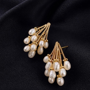 May include: A pair of gold-colored earrings with a cluster of white, teardrop-shaped pearls. The earrings have a delicate, fan-like design, with the pearls suspended from a gold framework. The background is a dark, textured fabric.