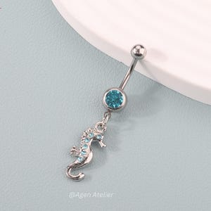 Puede incluir: Un piercing para el ombligo plateado con una gema turquesa y un dije de caballito de mar. El caballito de mar está detallado con pequeñas gemas turquesas. El piercing se muestra sobre una superficie azul claro.