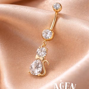 May include: Gold-toned belly button ring featuring a cat design with clear, sparkling cubic zirconia stones. The cat pendant includes a pear-shaped stone and a round stone for the head. The ring is curved and has a round stone at the top.