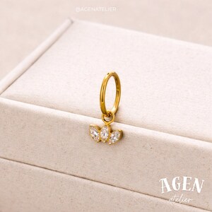 May include: A gold-colored hoop earring with a dangling charm. The charm features three clear, sparkling gemstones arranged in a floral or leaf-like pattern. The earring is set against a neutral, off-white background.