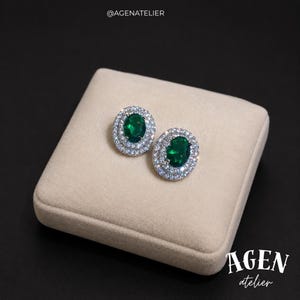 May include: A pair of oval-shaped stud earrings featuring a central deep green gemstone surrounded by a double halo of small clear white stones. The text @AGENATELIER appears at the top and AGEN atelier is printed at the bottom right corner.