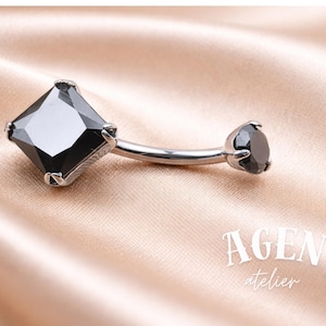 May include: A curved belly button ring with a silver-colored bar. The ring features a large, square, black gemstone on one end and a smaller, round, black gemstone on the other. The background is a satin fabric.