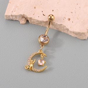 May include: Gold-coloured belly button ring with a clear gem at the top and a dangling charm. The charm features a crescent moon with a cat silhouette and a star pendant, all adorned with small, clear crystals. The jewellery is set against a light blue background.