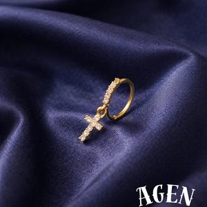 May include: A gold earring featuring a cross pendant, embellished with tiny, glittering stones. The earring is displayed against a backdrop of deep blue, satin-textured fabric. The text "AGEN atelier" is visible.