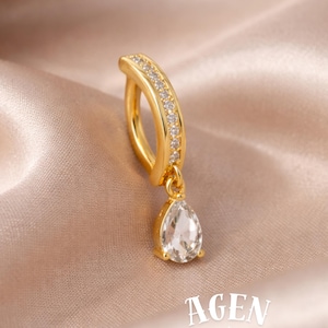 May include: A gold-colored belly button ring with a curved bar, adorned with small, clear gemstones. A teardrop-shaped, clear gemstone hangs from the bar. The word "AGEN" and "atelier" are printed on the bottom right.