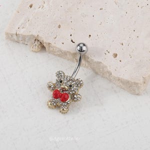May include: A silver belly button ring featuring a sparkling teddy bear design. The bear is adorned with clear crystals and a red bow tie. The jewelry is set against a neutral background.