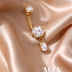May include: A gold-colored belly button ring with three clear, sparkling cubic zirconia stones. The curved bar has a small stone at the top, a larger round stone in the middle, and a teardrop-shaped stone dangling below. The word "AGEN atelier" is in the image.