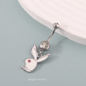 May include: A silver-colored belly button ring featuring a white bunny design with a pink gem eye. The ring has a curved barbell with a clear gem and a small ball at the top. The design is on a light blue surface.