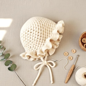 May include: A cream-colored, crocheted baby bonnet with a ruffled brim and tie closure. A small basket holds several wooden buttons. A crochet hook and a ball of yarn are also present, suggesting a handmade item.