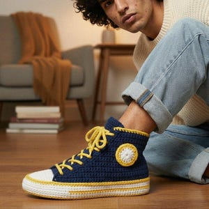 May include: A navy blue and white high-top sneaker with yellow detailing. The shoe is crocheted with a white toe cap, a yellow sole, and a yellow star-shaped patch. The laces are yellow.