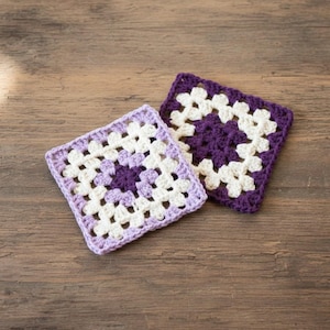 May include: Two handmade crochet squares in a granny square pattern. One is lavender, white, and purple, the other is purple and white. The squares are on a wooden surface. The squares are likely coasters or decorative items.