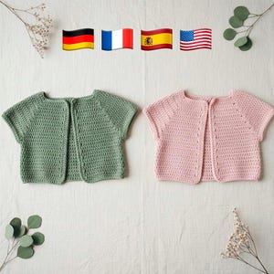 Op de afbeelding: Twee handgemaakte gehaakte babyvestjes. Eén is saliegroen, de andere lichtroze. Beide hebben korte mouwen en een knoopsluiting. De vestjes worden gepresenteerd op een witte ondergrond met decoratief groen.