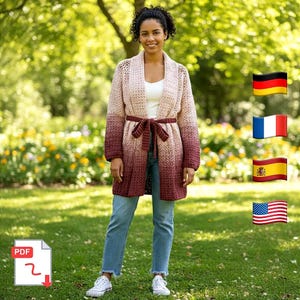 May include: A long, open-front crochet cardigan in a gradient of colors from light pink to burgundy. The cardigan has long sleeves and a matching belt. The person is wearing blue jeans and white sneakers. The background is a green, grassy area.