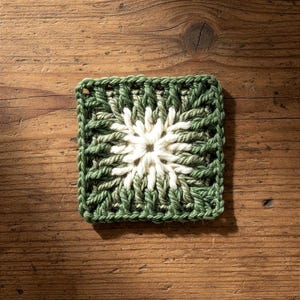 Può includere: Sottobicchiere quadrato fatto a mano all'uncinetto, nei toni del verde e del bianco. Il sottobicchiere presenta un motivo floreale bianco centrale circondato da un bordo verde strutturato. Posizionato su una superficie di legno.
