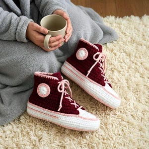 Op de afbeelding: Een paar bordeauxrode en witte gehaakte hoge slippers met roze accenten. De slippers hebben een witte neus, roze veters en een cirkelvormige patch aan de zijkant. Ze liggen op een crèmekleurig tapijt.