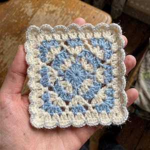 May include: A square, handmade crochet doily in cream and light blue. The center features a blue floral design, surrounded by cream-colored crochet. The edges are scalloped and trimmed with silver thread. This decorative piece is perfect for home decor.