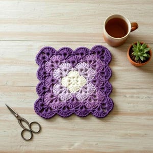 Könnte beinhalten: Ein handgefertigter, quadratischer Häkeluntersetzer in Lila- und Weißtönen liegt auf einer hellen Holzoberfläche. Eine Tasse Tee, eine kleine Topf-Sukkulente und eine Schere befinden sich ebenfalls auf dem Tisch. Der Untersetzer hat einen gewellten Rand.