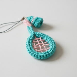 Puede incluir: Un charm de raqueta de tenis de ganchillo hecho a mano en turquesa, rosa y blanco. La cabeza de la raqueta es rosa con un borde turquesa, y el mango es turquesa y rosa. Se adjunta una cuerda negra para colgar.