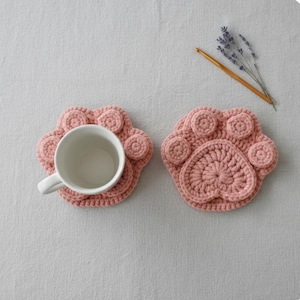 Może przedstawiać: Dwa różowe, szydełkowe podkładki w kształcie łapy. Jedna podkładka trzyma biały kubek. Podkładki wykonane są z miękkiej, teksturowanej przędzy. Projekt przedstawia odcisk łapy z okrągłymi poduszkami.