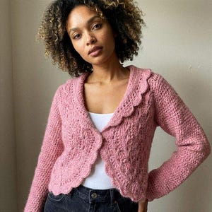 Puede incluir: Un cárdigan rosa de ganchillo con cuello festoneado y cierre de botón. El suéter de manga larga tiene un patrón de puntada texturizada. La prenda se usa sobre una camiseta blanca y se combina con jeans oscuros.